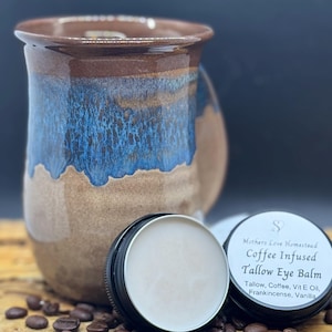 May include: A ceramic mug with brown and blue glaze sits next to two small tins of coffee-infused tallow eye balm. The tins are open, revealing the balm. Coffee beans are scattered around the tins.