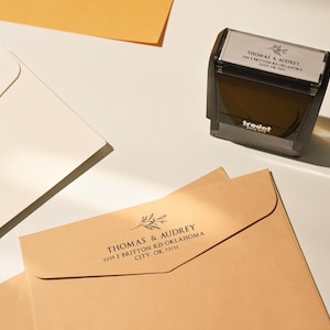 May include: A black Trodat stamp with the text "THOMAS & AUDREY" and address information. Several cream and tan envelopes are arranged around the stamp. A yellow paper is in the background.