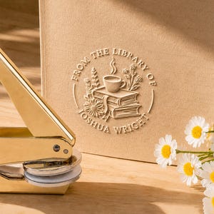 Könnte beinhalten: Ein goldfarbenes Metall-Prägewerkzeug neben einer beigen Papieroberfläche mit einem kreisförmigen Prägemotiv. Das Design zeigt einen Bücherstapel, eine dampfende Tasse und Blumen sowie den Text FROM THE LIBRARY OF JOSHUA WRIGHT.
