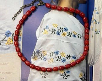 May include: A red coral necklace with a silver clasp. The necklace is made of small, round beads and is lying on a white fabric with embroidered flowers.