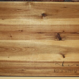 May include: A wooden chopping board with a brown leather handle. The board has a natural wood grain and is engraved with the text "home" and coordinates.