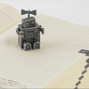 May include: A small, metallic robot figurine with the words "YOU LOVE YOU" on its chest sits atop an open notebook. The robot has a vintage, industrial design with a bow-tie-shaped antenna. The notebook has a grid pattern and a brown spine.