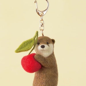 Könnte beinhalten: Ein braunes Filzotter, das einen roten Filzapfel mit einem grünen Filzblatt hält. Das Otter ist an einem goldenen Schlüsselanhänger befestigt.