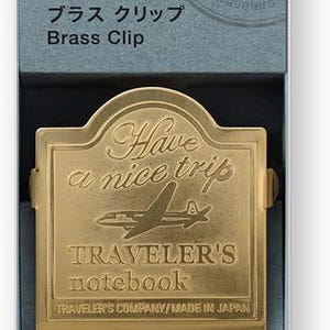 May include: A brass clip for a Traveler's notebook, presented in a blue box. The clip is gold-colored, with the text "Have a nice trip" and a plane design. The box also displays the text "TRAVELER'S notebook".