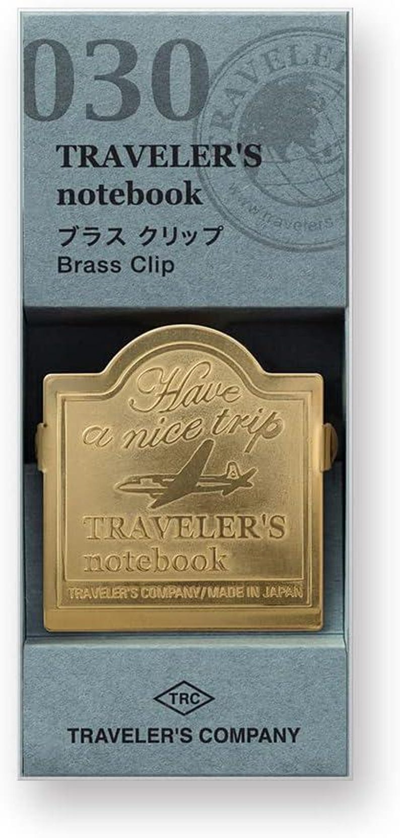 May include: A brass clip for a Traveler's notebook, presented in a blue box. The clip is gold-colored, with the text "Have a nice trip" and a plane design. The box also displays the text "TRAVELER'S notebook".
