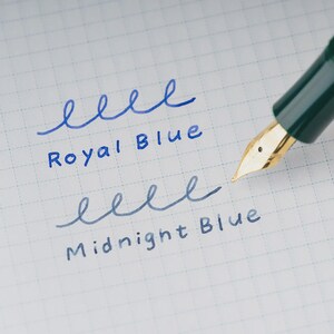Puede incluir: Primer plano de una pluma estilogr&aacute;fica con plum&iacute;n dorado, escribiendo sobre papel cuadriculado. La pluma escribe las palabras "Royal Blue" y "Midnight Blue" en tinta azul, mostrando los colores de la tinta.