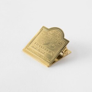 May include: A gold-colored metal clip with the text "Have a nice trip" and "Traveler's Notebook" engraved on it. The clip has a rectangular shape with a curved top and a hinged design, perfect for securing pages.
