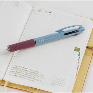 Può includere: Una penna multi-colore blu e bordeaux è appoggiata su un quaderno aperto. La penna ha il testo "Hobonichi Techo 2023" stampato sul fusto. Il quaderno è aperto su una pagina a griglia con la data "15". Il quaderno ha una copertina gialla.