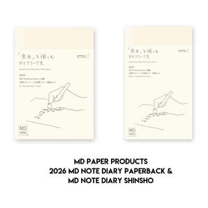 May include: Two cream-coloured MD Paper Products notebooks. The notebooks have Japanese text and illustrations of a hand writing with a pen. The text "2026 MD NOTE DIARY PAPERBACK & MD NOTE DIARY SHINSHO" is below the notebooks.