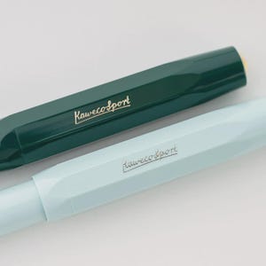 Puede incluir: Primer plano de dos bol&iacute;grafos Kaweco Sport. Uno es de color verde azulado oscuro y el otro azul pastel claro. Ambos bol&iacute;grafos tienen el logotipo de Kaweco Sport en letras doradas. Los bol&iacute;grafos est&aacute;n en &aacute;ngulo, mostrando su dise&ntilde;o octogonal.