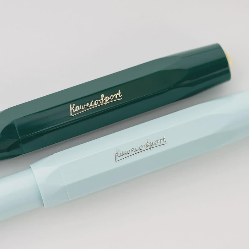 Puede incluir: Primer plano de dos bol&iacute;grafos Kaweco Sport. Uno es de color verde azulado oscuro y el otro azul pastel claro. Ambos bol&iacute;grafos tienen el logotipo de Kaweco Sport en letras doradas. Los bol&iacute;grafos est&aacute;n en &aacute;ngulo, mostrando su dise&ntilde;o octogonal.