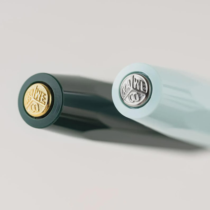 Puede incluir: Primer plano de dos plumas estilogr&aacute;ficas. Una pluma es de color verde oscuro con un emblema dorado, y la otra es azul claro con un emblema plateado. Ambas plumas tienen la marca "KAWECO" en el extremo.