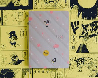 May include: A 2026 planner with a gray cover featuring a diagonal striped pattern, pink accents, and cartoon illustrations. The planner is set against a backdrop of yellow comic book panels with Japanese text.