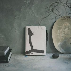 Puede incluir: Un diario blanco con un gran diseño de pincelada negra en forma de "L". Un bolígrafo negro está sujeto al lateral. El diario está sobre una superficie gris junto a una pila de libros y un plato decorativo.