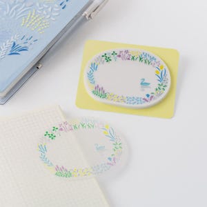 May include: Oval-shaped sticky notes with floral and duck designs. One is on a yellow pad, the other is transparent. A light blue notebook and pen are in the background. The notes have a white center with a duck illustration.
