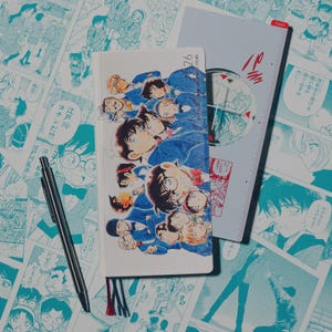 Może przedstawiać: Biały notatnik z kolorową ilustracją anime przedstawiającą wiele postaci w niebieskich mundurach. Widoczny jest również srebrny długopis i szara linijka z japońskim tekstem. Tło to strona komiksu.