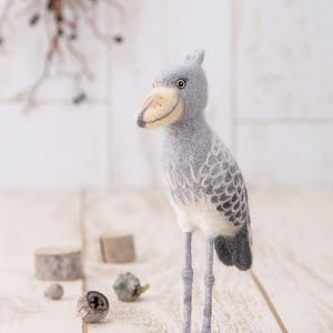 Op de afbeelding: Een grijs en witte naaldgevoelde vogel met een lange snavel en lange poten. De vogel staat op een wit houten oppervlak.