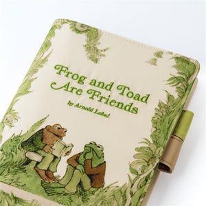 Puede incluir: Una portada de libro con el título "Frog and Toad Are Friends" de Arnold Lobel. La portada tiene un fondo crema con ilustraciones verdes de follaje y los dos personajes. Rana lee un libro a Sapo.