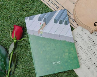 May include: A green 2026 planner with a piano keys illustration and a small figure holding a rose. A red rose and sheet music are next to the planner on a bed of green grass.
