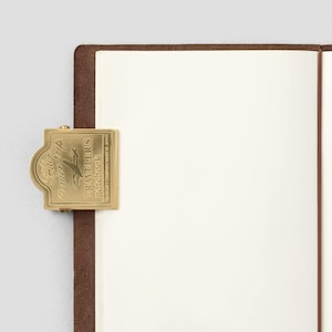 May include: A brass-colored metal clip with the text "Have a nice trip" and "TRAVELER'S notebook" is attached to a brown leather notebook. The notebook is open, revealing blank white pages. The clip features an airplane graphic.