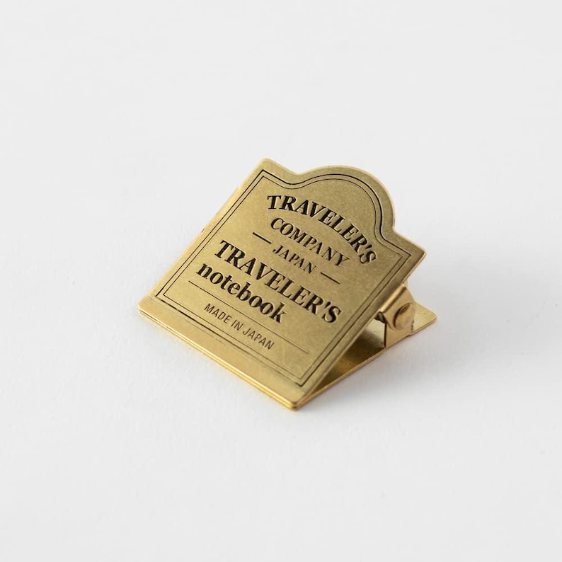 May include: A brass-colored metal clip with the text "TRAVELER'S COMPANY JAPAN TRAVELER'S notebook MADE IN JAPAN". The clip has a rectangular shape with a rounded top and a hinged clasp.