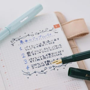 Puede incluir: Una pluma estilogr&aacute;fica azul claro y una pluma estilogr&aacute;fica verde oscuro est&aacute;n sobre un cuaderno con texto japon&eacute;s escrito a mano. El texto describe los pasos para hacer una tarta de manzana. El cuaderno est&aacute; abierto en una p&aacute;gina con un calendario.