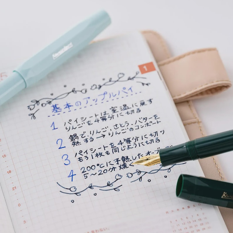 Puede incluir: Una pluma estilogr&aacute;fica azul claro y una pluma estilogr&aacute;fica verde oscuro est&aacute;n sobre un cuaderno con texto japon&eacute;s escrito a mano. El texto describe los pasos para hacer una tarta de manzana. El cuaderno est&aacute; abierto en una p&aacute;gina con un calendario.