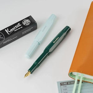 Puede incluir: Dos plumas estilogr&aacute;ficas, una azul claro y otra verde oscuro, se muestran con una caja negra y un cuaderno marr&oacute;n. La pluma verde lleva la etiqueta "Kaweco Sport". La caja tiene el logotipo de Kaweco y el texto "GERMANY, SINCE 1883". Se ve un peque&ntilde;o horario.