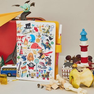 Puede incluir: Un cuaderno colorido con una cubierta amarilla y un portalápices, con pegatinas de dibujos animados caprichosos. Un faro azul y rojo, un autobús en miniatura y un sujetalibros con forma de pájaro también son visibles. Una manzana parcialmente comida y uvas están en primer plano.