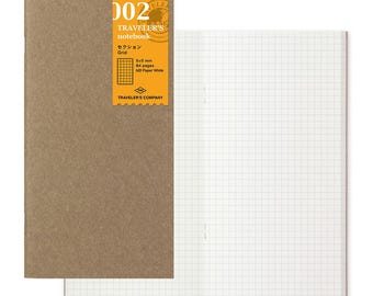 May include: A Traveler's Company notebook with a brown cover and a gridded page. The cover has a yellow label with the text "002 Traveler's notebook Grid 5x5 mm 64 pages MD Paper White Traveler's Company".