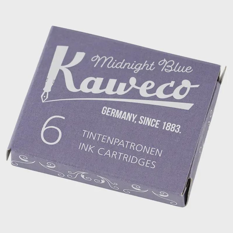 Puede incluir: Una caja de cartuchos de tinta Kaweco Midnight Blue. La caja es de color morado apagado con texto blanco que dice "Kaweco", "Midnight Blue", "Germany, Since 1883" y "6 Ink Cartridges". La caja tiene remolinos decorativos.