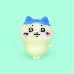 Könnte beinhalten: Eine kleine, weiße Filzkatze mit blauen Ohren und einem blauen Stirnband. Die Katze hat schwarze Augen, eine rosa Nase und einen rosa Rouge auf den Wangen. Sie steht auf einem hellblauen Hintergrund.