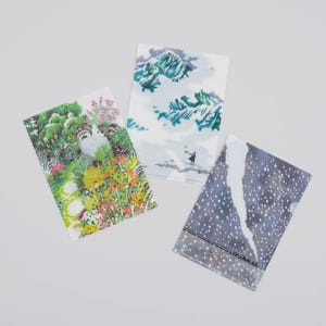 Puede incluir: Tres carpetas de papel decorativas con ilustraciones de temática natural. Una presenta un búho en un jardín, otra representa montañas y la tercera un paisaje nevado. Las carpetas están sobre un fondo blanco.