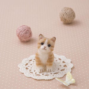 Puede incluir: Una pequeña figurita de gatito de fieltro naranja y blanco se sienta sobre un encaje de ganchillo blanco. El gatito tiene una cara blanca con marcas naranjas y mira directamente a la cámara. Hay una bola de lana rosa y una bola de lana beige en el fondo.
