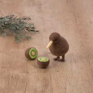 Può includere: Una statuetta di uccello kiwi in feltro marrone con un lungo becco, in piedi su una superficie di legno con due kiwi tagliati a metà accanto.