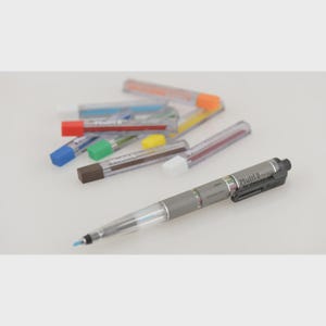 May include: A gray and clear Multi 8 Pilot pen with a black clip and several colored lead refills. The pen has the text "Multi 8 Pilot" and "for checking use" printed on the side. The lead refills have various colored tips.