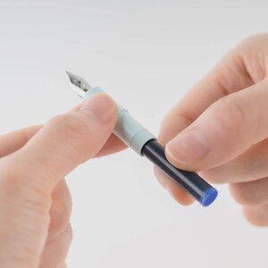 Puede incluir: Una pluma estilogr&aacute;fica con un cuerpo azul claro y un cartucho de tinta azul oscuro. La pluma es plateada y el bol&iacute;grafo se sostiene entre dos manos sobre un fondo blanco.