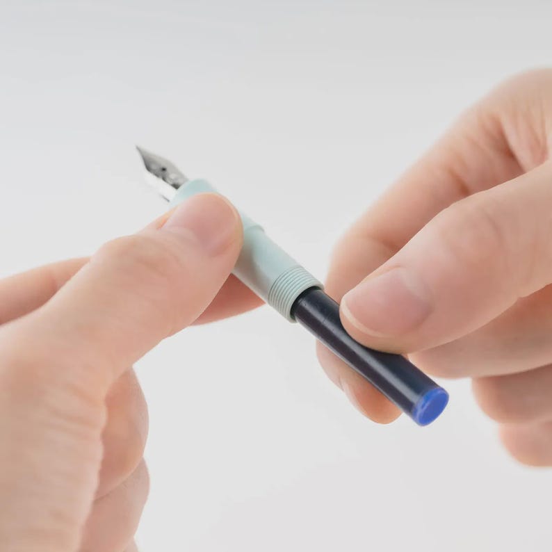 Puede incluir: Una pluma estilogr&aacute;fica con un cuerpo azul claro y un cartucho de tinta azul oscuro. La pluma es plateada y el bol&iacute;grafo se sostiene entre dos manos sobre un fondo blanco.