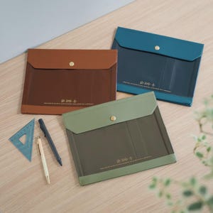 Puede incluir: Tres fundas para documentos en marrón, verde azulado y verde oliva con un cuerpo transparente y una solapa de color liso asegurada con un broche dorado. Una regla triangular azul y dos bolígrafos están al lado. El texto en las fundas dice: "Escribir a mano es como enviar tu mente de viaje."