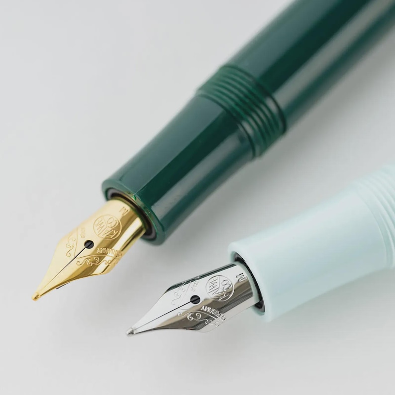 Puede incluir: Primer plano de dos plumas estilogr&aacute;ficas. Una es verde oscuro con un plum&iacute;n dorado, la otra es azul claro con un plum&iacute;n plateado. Ambas plumas tienen un dise&ntilde;o elegante y est&aacute;n sobre un fondo blanco.