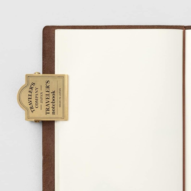 May include: A brass notebook clasp from Traveler's Company Japan, attached to a brown leather notebook. The clasp is rectangular with rounded edges and features the brand name and product information. The notebook pages are white and blank.