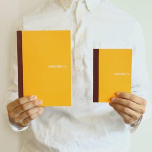 May include: Two yellow notebooks with brown spines are held by a person wearing a white shirt. The notebooks have text in Japanese. The larger notebook is rectangular, and the smaller one is square.