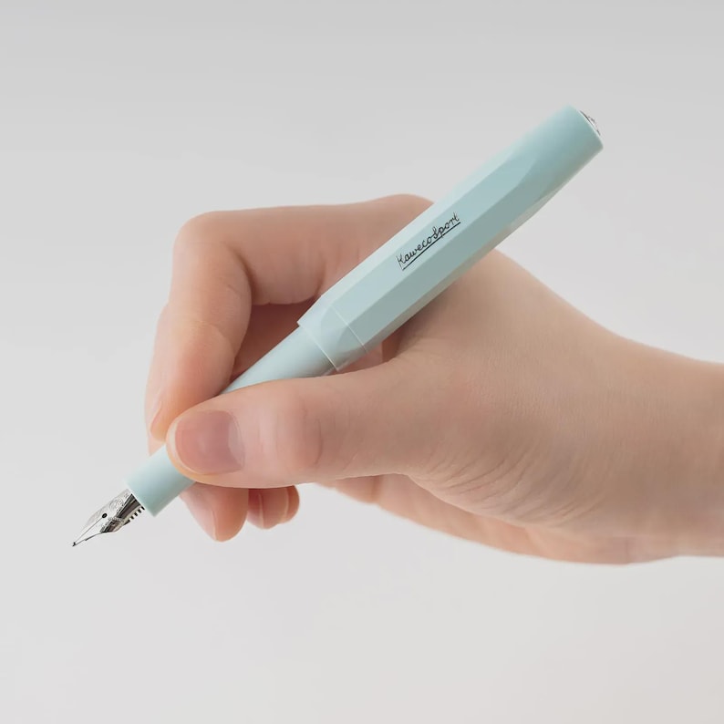 Puede incluir: Una pluma estilogr&aacute;fica Kaweco Sport azul claro. El bol&iacute;grafo se sostiene en una mano y tiene el nombre de la marca impreso en el lateral. El bol&iacute;grafo tiene un plum&iacute;n plateado y un capuch&oacute;n. El fondo es blanco.