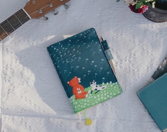 May include: A teal-colored journal with a whimsical illustration of a red bear and a black and white cat on a green field. The journal has a pen inserted and a tassel. A ukulele and flowers are in the background.