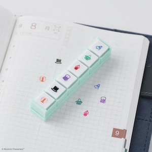 Puede incluir: Un juego de sellos rectangular verde menta con siete sellos cuadrados blancos con ilustraciones coloridas. Los sellos están dispuestos en un diseño escalonado y descansan sobre un cuaderno abierto. Las páginas del cuaderno tienen un patrón de cuadrícula.