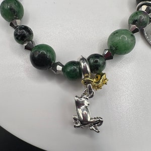 May include: A green and black gemstone bracelet with a silver frog charm. The bracelet is made with round beads and has a gold accent on the charm.