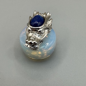 May include: A silver dragon head pendant with a blue gemstone set on a round, opalescent stone. The dragon's head has detailed features, and the opalescent stone has a soft, glowing appearance. The pendant is likely a piece of jewellery.