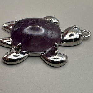 Puede incluir: Un amuleto de tortuga de plata con una piedra de amatista púrpura en el centro.