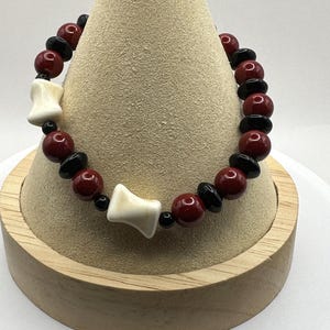 May include: A red and black beaded bracelet with white bone-like charms. The bracelet is displayed on a wooden stand.