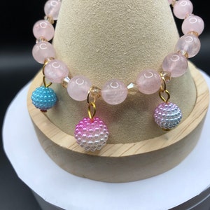 Puede incluir: Un brazalete de cuarzo rosa con tres abalorios de cuentas en rosa, azul y blanco.
